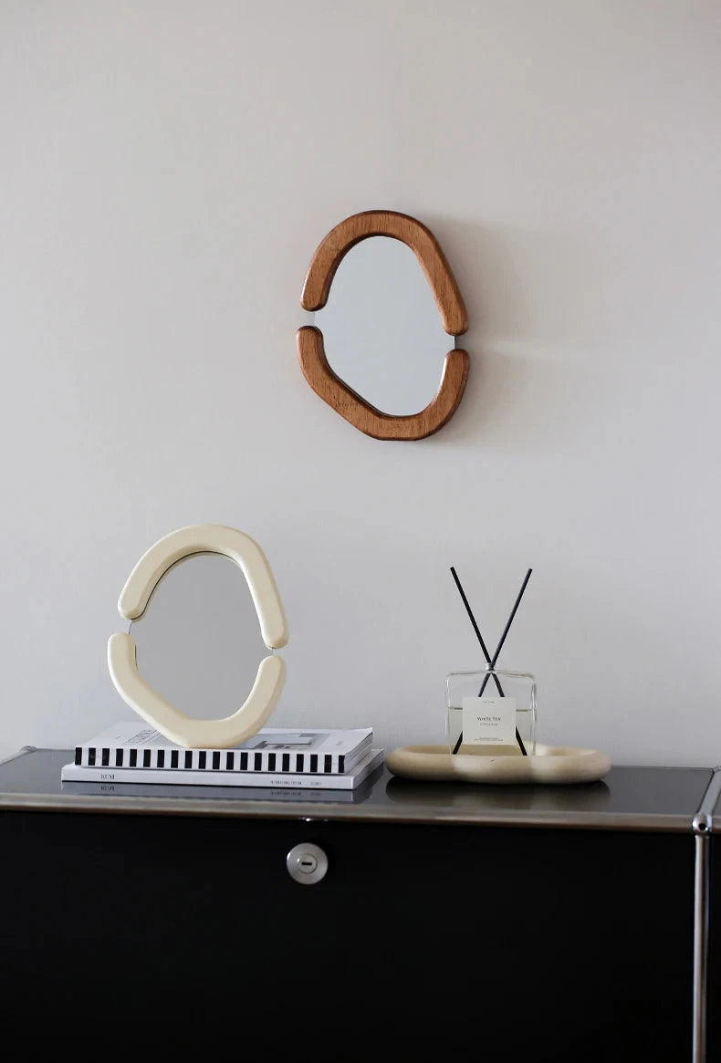 A beautifully crafted Zia Mirror showcasing its retro design and organic shape. The mirror features a smooth wooden frame available in both wood and cream finishes, adding a touch of elegance and warmth. It is displayed both as a countertop mirror and mounted on a wall, demonstrating its versatile use. This mirror serves as a stylish vanity accessory and a chic decor accent, perfect for home décor enthusiasts looking to enhance their space with unique, sophisticated pieces.