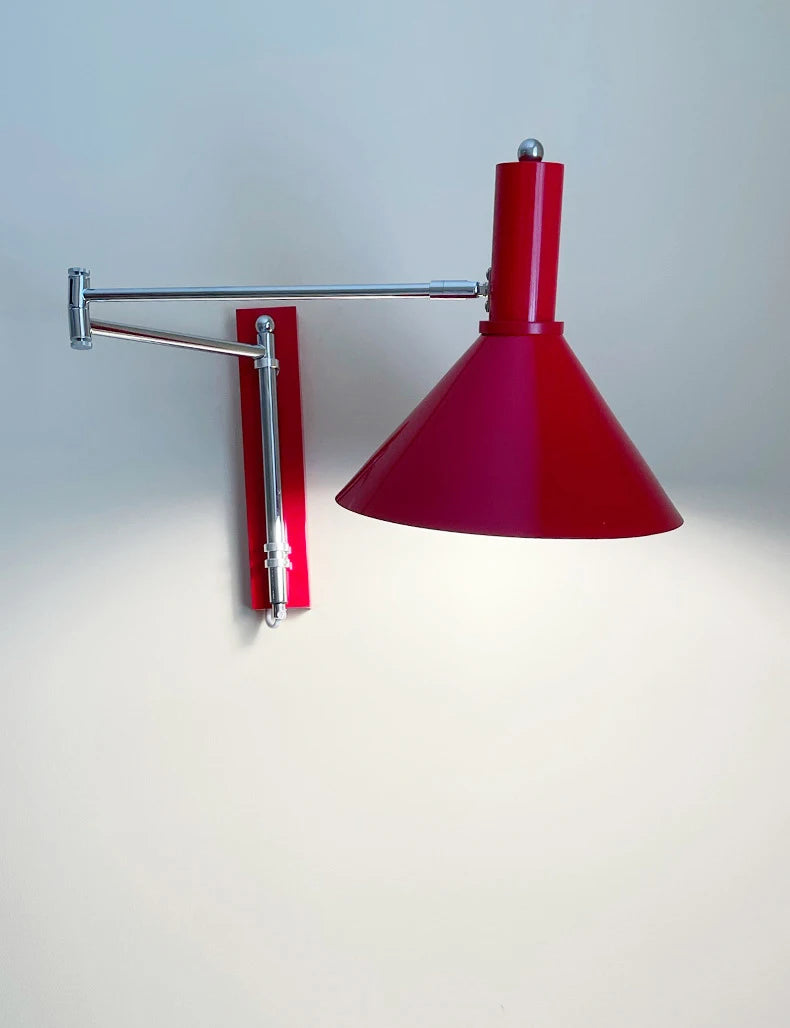 A Colleen Wall Lamp featuring a sleek, modern design with silver hardware. The lamp includes a moveable arm for adjustable lighting, ideal for bedside reading or accent lighting. Available in vibrant red or classic white, it can be installed with or without a plug cable. The lamp is displayed in a stylish home setting, highlighting its versatility and appeal for home decor enthusiasts looking to enhance their space with both functionality and style.