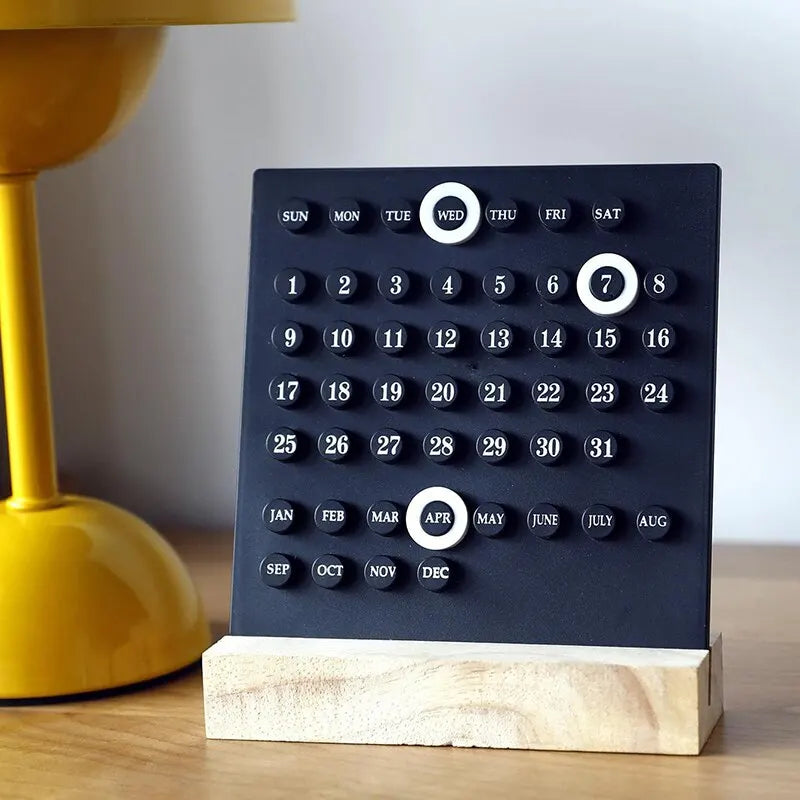 Image of the Callie Calendar, a compact, reusable calendar in black or white with a sleek acrylic design and a natural wooden stand. Features a red marker for highlighting important dates, perfect for organizing spaces like the kitchen or office with a touch of style.