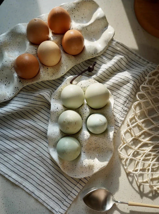 A ceramic Umiko Egg Crate featuring an organic shape, ivory finish with black paint splatters, available in half a dozen or a dozen slots, adding uniqueness to kitchen décor.