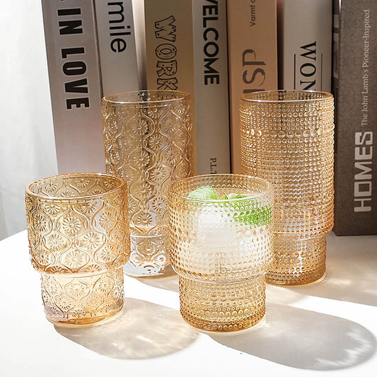 Image of the Unai Glass Collection featuring elegant tumbler and highball glasses. Made from laminated glass with intricate beaded or floral designs, these stackable glasses come in amber or clear colors. Available in two-piece or six-piece sets, perfect for entertaining and enhancing your home decor.